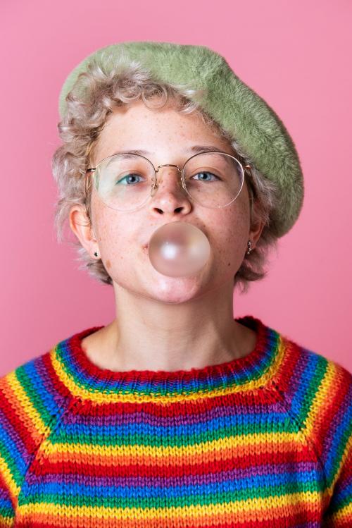 Cheerful girl wearing a green beret and a rainbow sweater chewing a bubble gum - 2223238